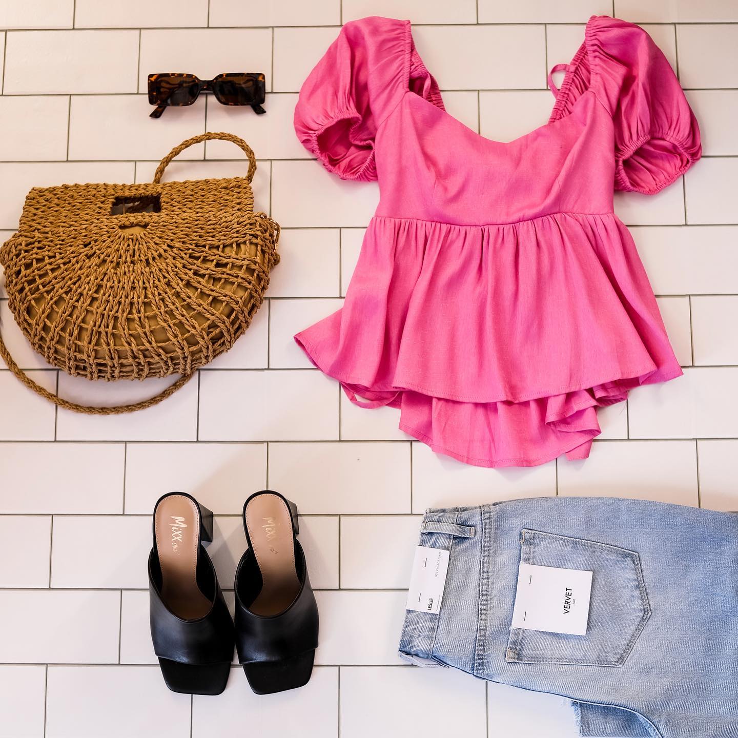 Flatlay of wicker purse, pink blouse, sunglasses, black heels, and and jeans